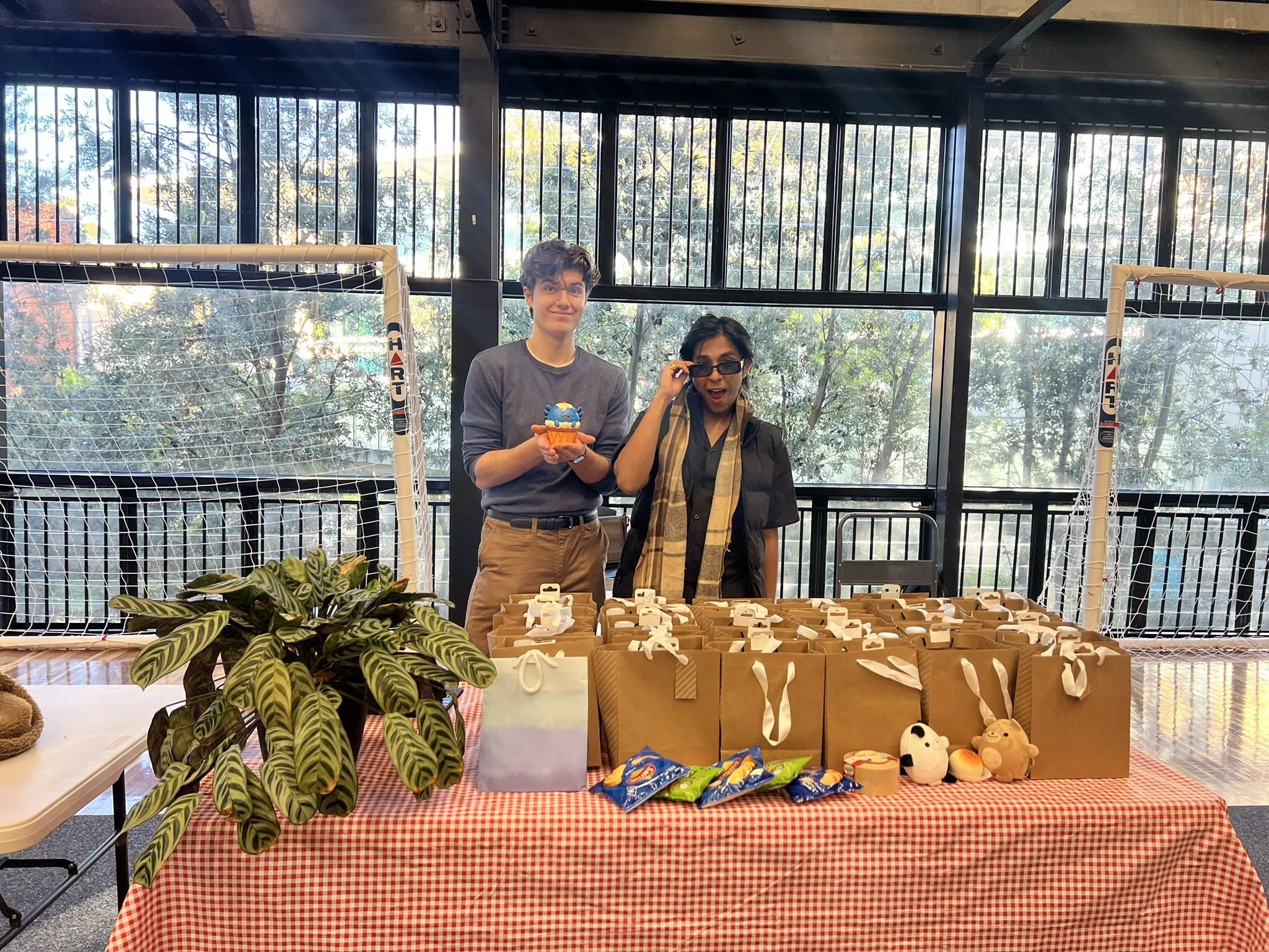 Two people standing behind a table at a craft fair, with one holding a cupcake-shaped plush toy. The table displays small gift boxes and toys and the background shows large windows with trees outside.