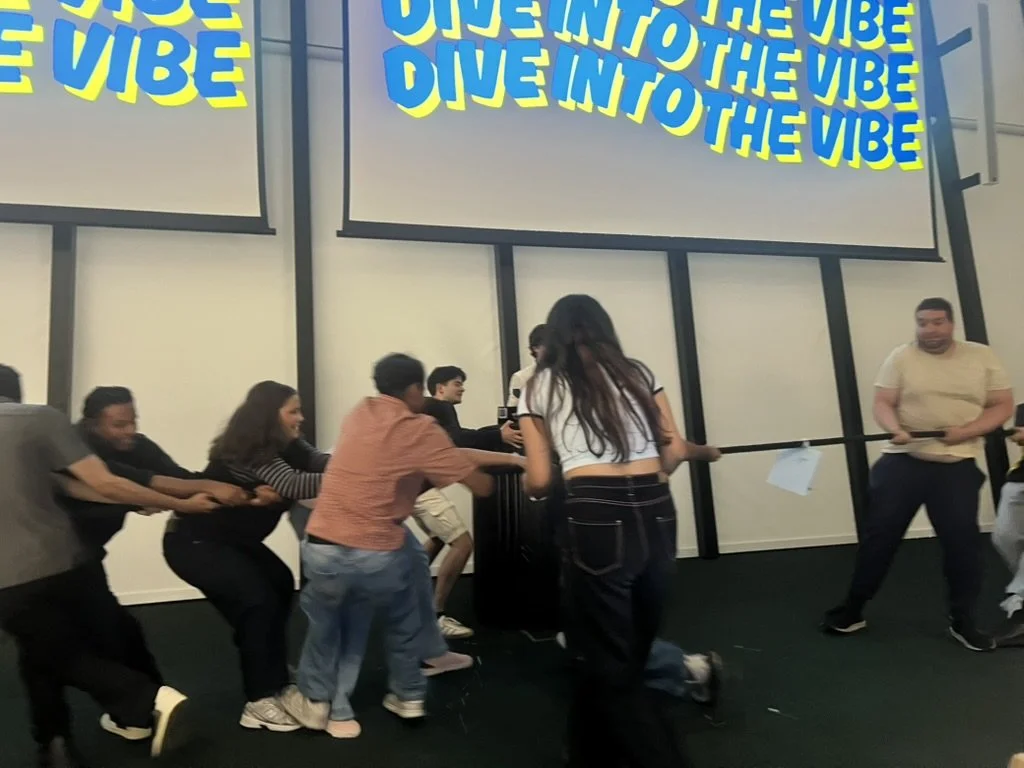 Group of people participating in a tug-of-war game indoors, with a large screen in the background displaying the text 'DIVE INTO THE VIBE' multiple times in colorful letters.