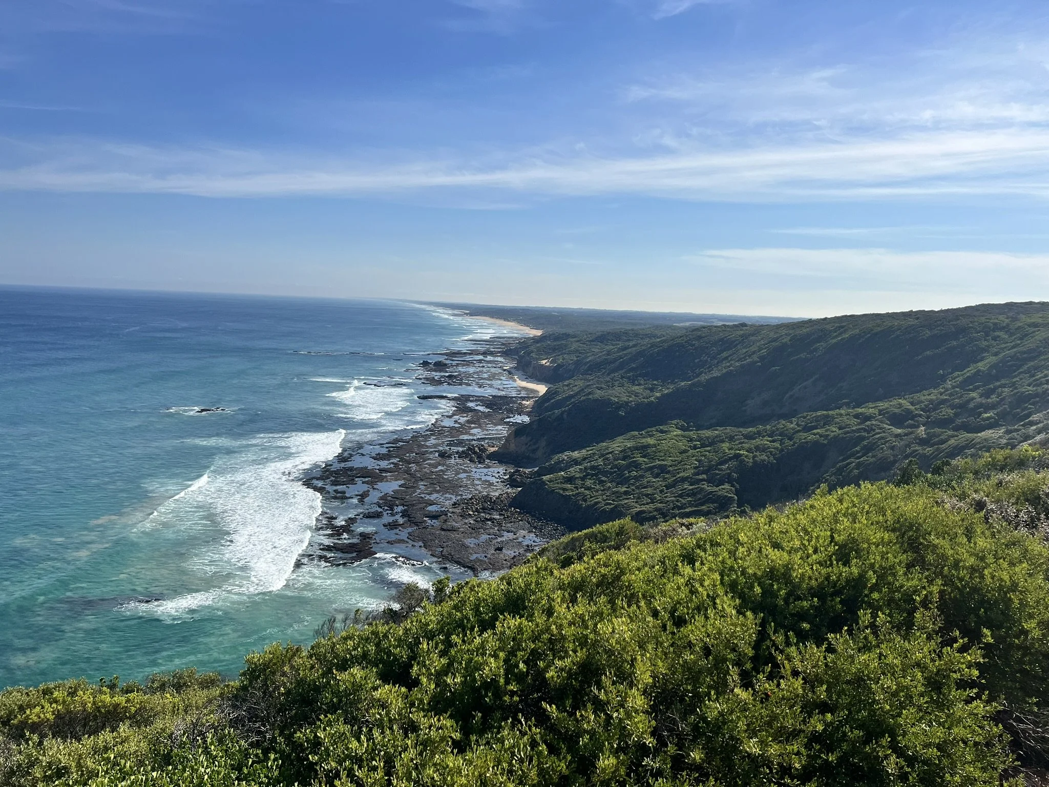 Coastline with ocean waves hitting rocks and a green hillside under a cloudy blue sky.