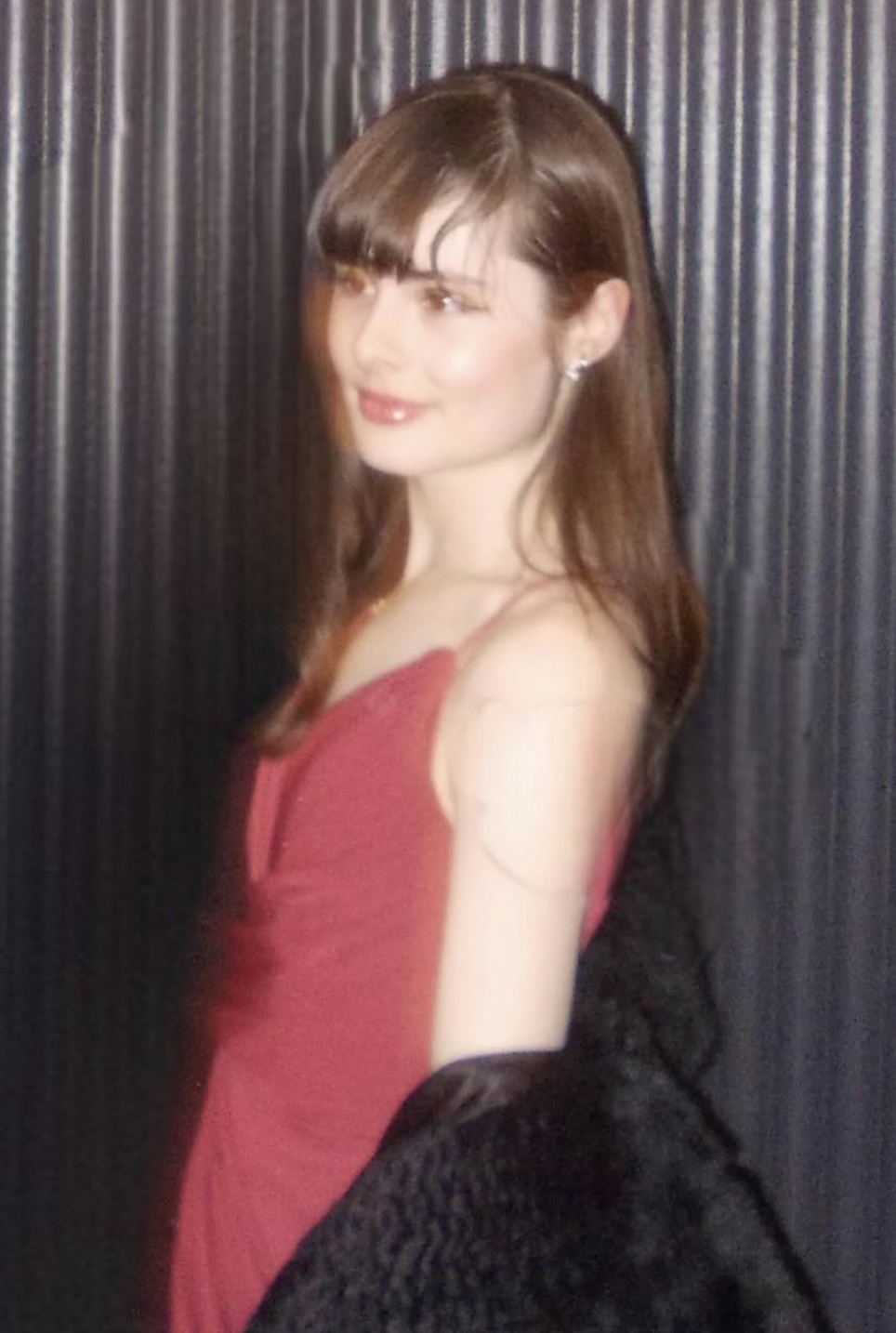 A young woman (Vice President) with long brown hair, wearing a red dress, standing in front of a dark, textured background.