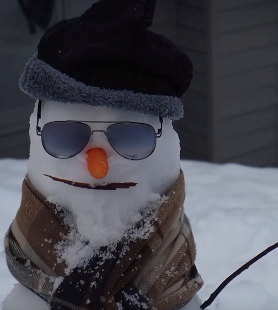 A snowman wearing sunglasses, a black hat with a furry gray band, a brown scarf, with a carrot nose, and a face with a drawn smile in a snowy outdoor setting.