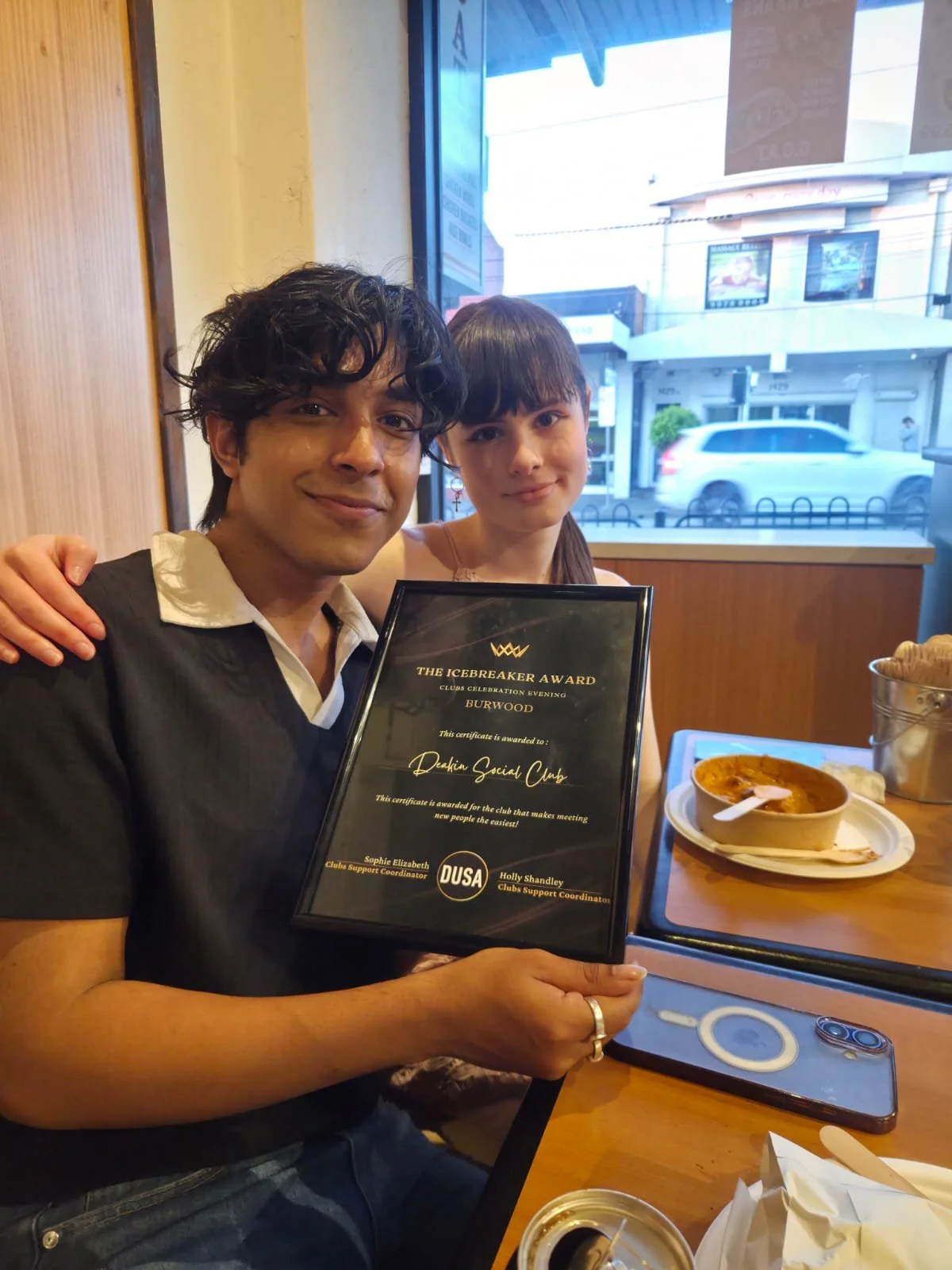 Two people sitting at a restaurant, smiling and holding a framed award certificate. The man on the left has dark wavy hair and is wearing a black shirt over a white polo. The woman on the right has straight dark hair with bangs and is wearing a light-colored top. There is food on the table, including a bowl of soup, bottles, and a smartphone.