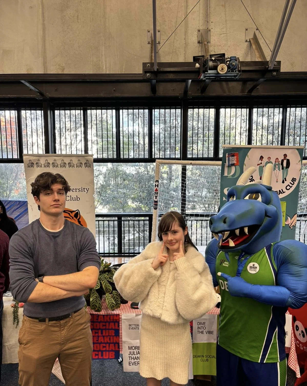 Three people posing inside a building with large windows, Deakin's dragon mascot in a green sports jersey, and various posters and banners behind them, with the words 'Deakin Social Club.'