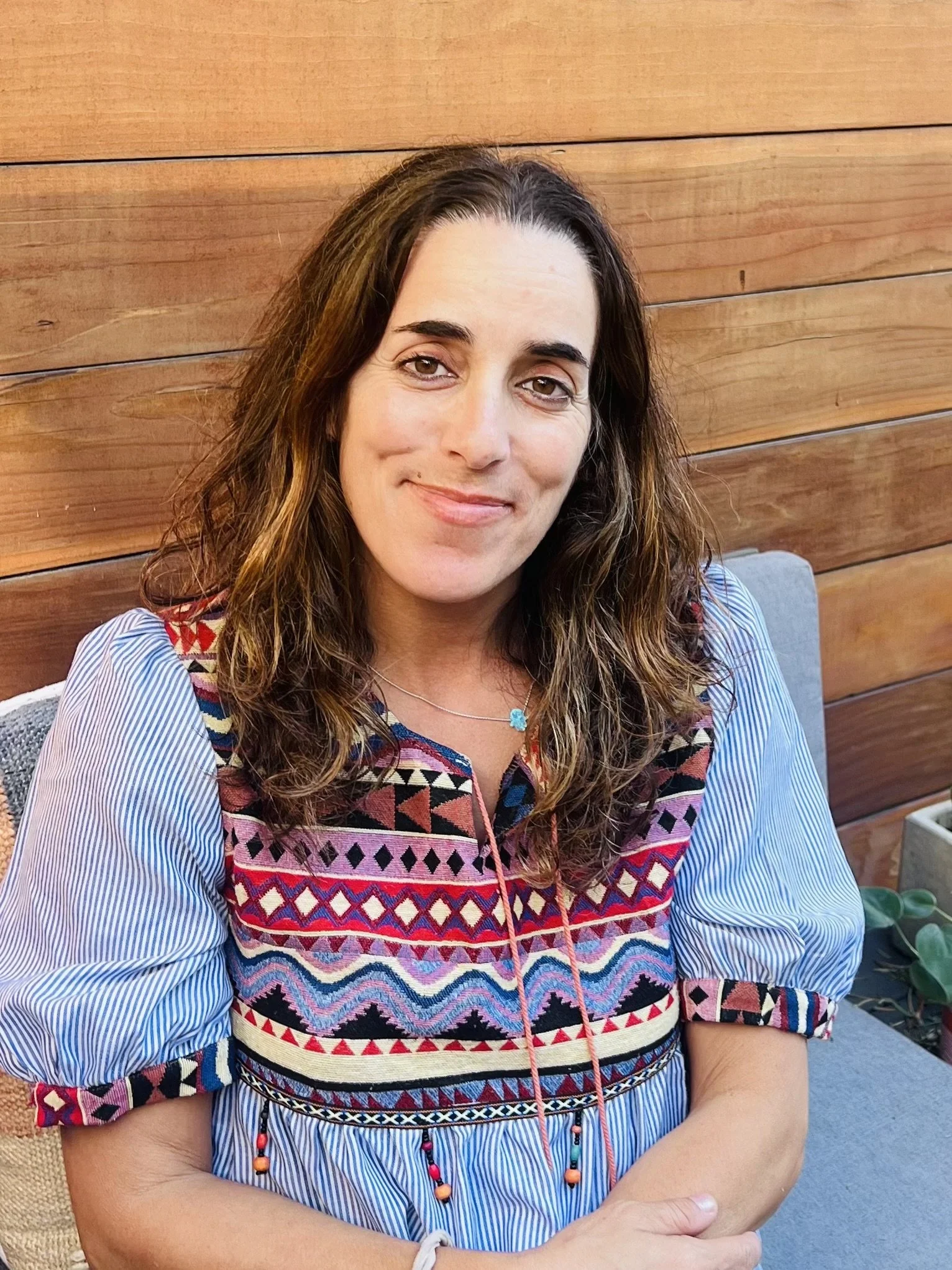 A woman with brown curly hair, light skin, and wearing a colorful embroidered top with striped sleeves, sitting outside against a wooden wall.