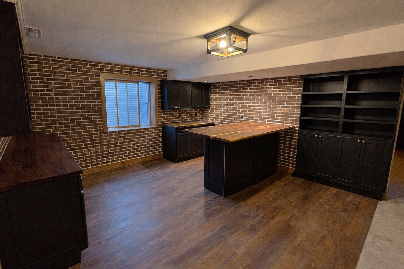 A basement kitchen with brick walls, wooden flooring, black cabinets, a wooden kitchen island, and a window with white shutters.