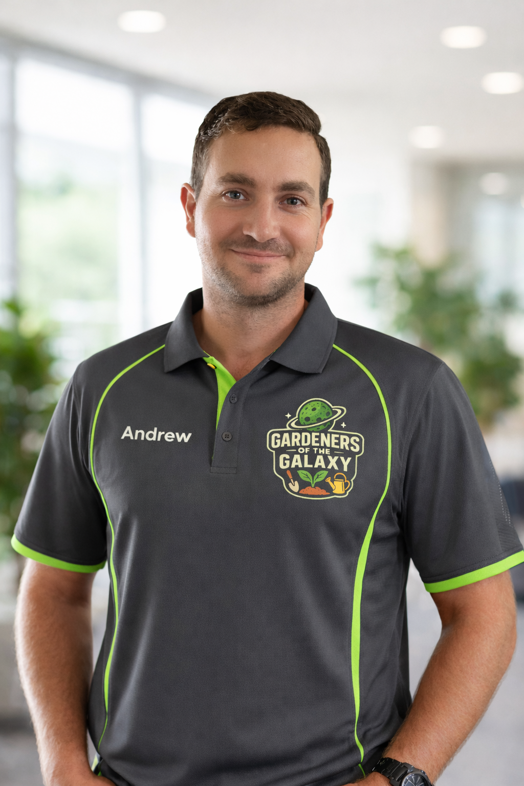 A man wearing a black shirt with green accents, labeled 'Andrew', stands indoors with a blurred background of windows and plants. His shirt has a logo for 'Gardeners of the Galaxy' featuring a planet with a ring, gardening tools, and a watering can.