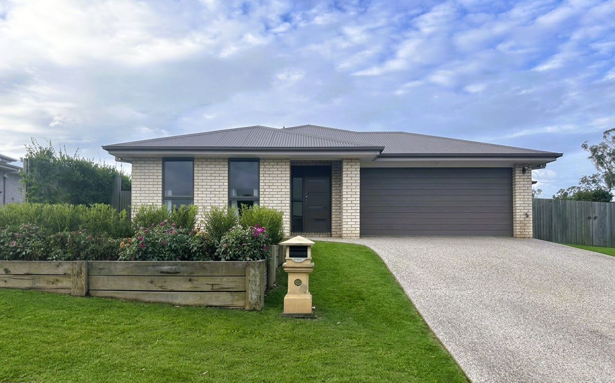 Front view of a modern house with a paved driveway, white brick exterior, three windows, a dark gray garage door, and a small front garden with green shrubs and flowers.