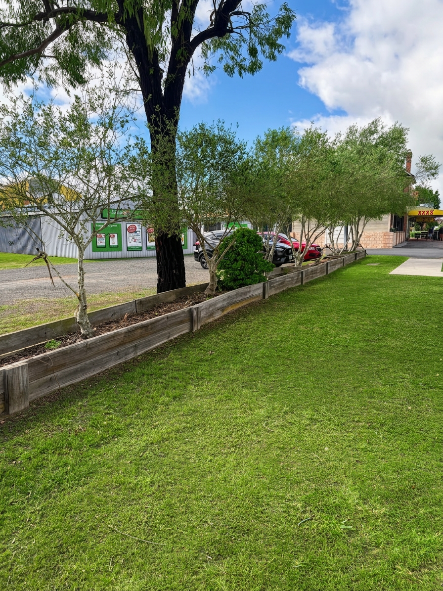 A grassy lawn with a row of trees and a wooden border, a parking lot with cars and a building with signage in the background, under a partly cloudy sky.