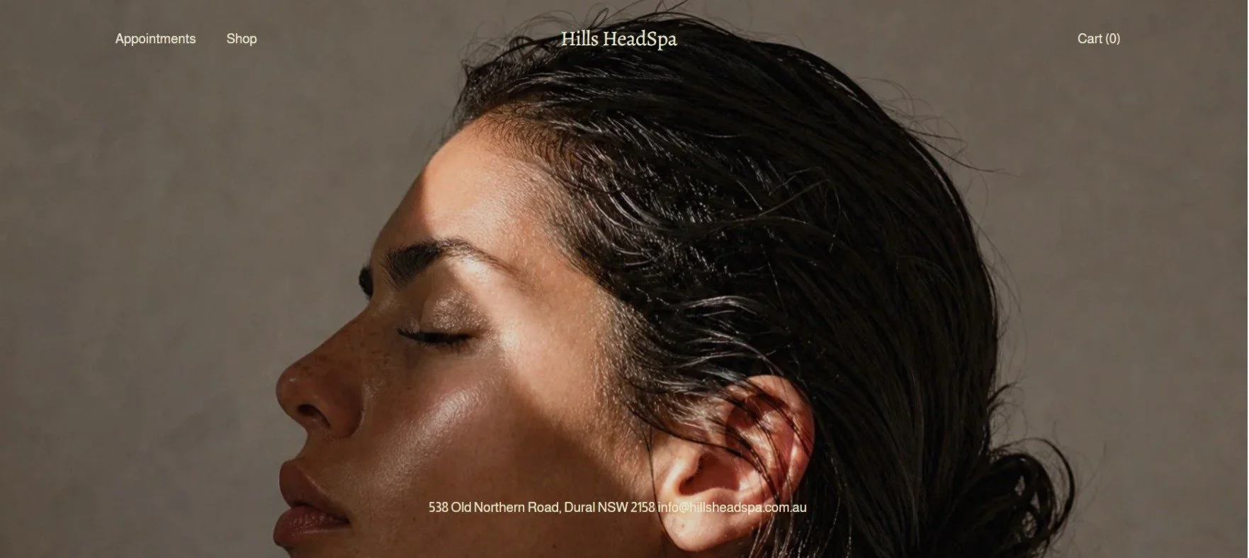 Close-up of a woman with wet, dark hair, her eyes closed, against a neutral background, promoting Hills Head Spa.