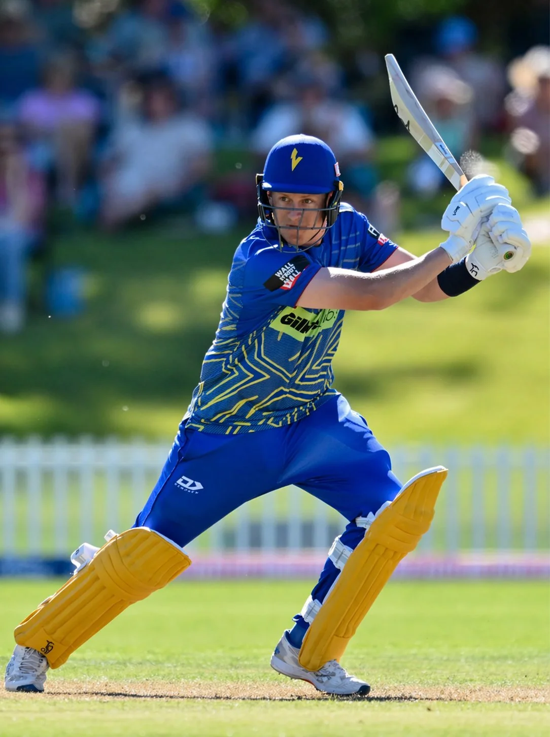 A cricket player in blue uniform and protective gear, including a helmet and pads, is batting on a cricket field, with spectators blurred in the background.