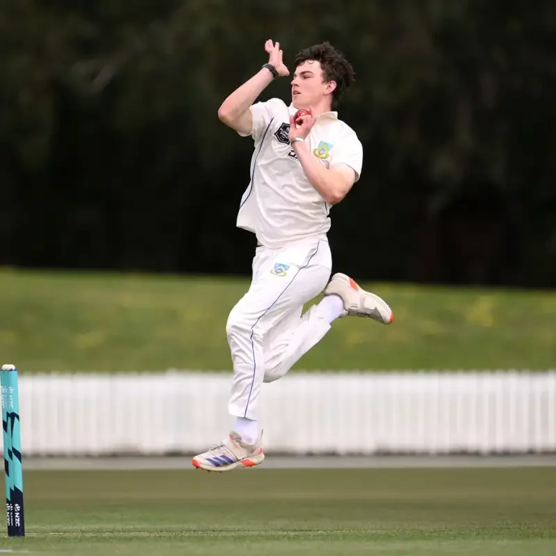 Cricket player jumping in the air, holding a red ball, on a cricket field.