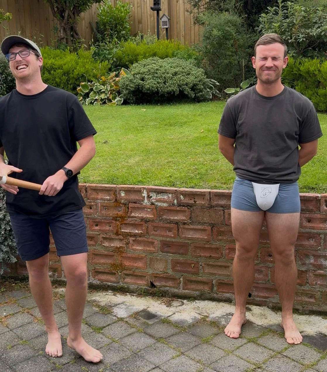 Two men standing outdoors on a brick patio with a green lawn and bushes behind them. The man on the left is holding an axe, wearing a black t-shirt, shorts, glasses, and a cap, and is laughing. The man on the right is standing with a slight smile, wearing a black t-shirt, gray shorts, and a white jockstrap, with his hands behind his back.