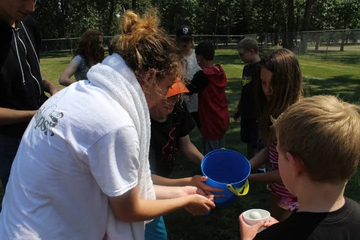 A group of children and an adult outside in a park, gathered around participating in an activity involving a blue bucket and cups.