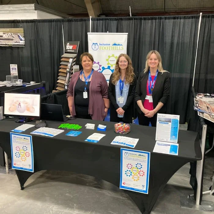 Three women standing behind an informational booth with various pamphlets, candy, and computer monitor, representing Inclusive Foothills at an indoor event.