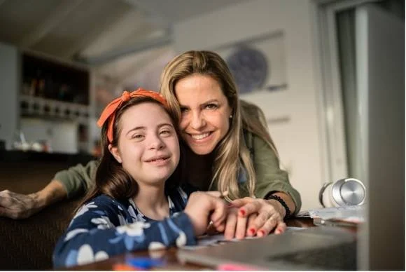 A woman and a girl smiling and sitting together at a table in a cozy kitchen. The girl wears an orange headband and a blue sweater with white flowers. The woman has long blonde hair and wears a green shirt. They are looking at a computer screen.