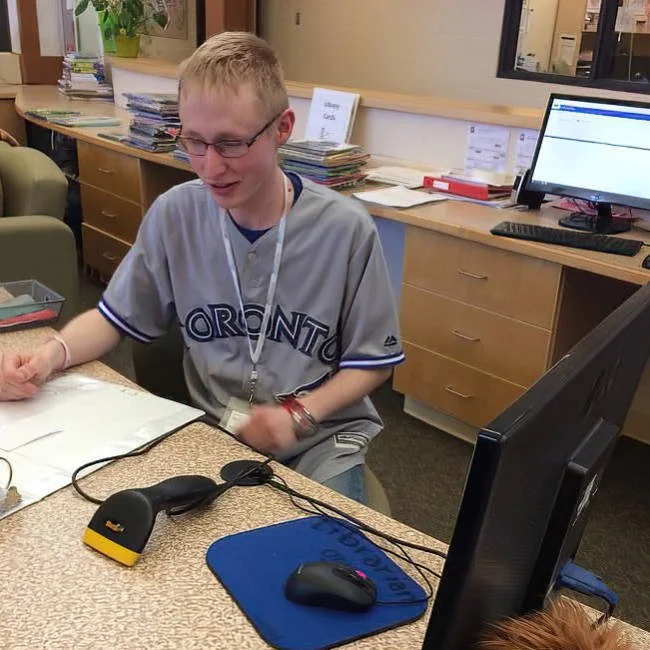 A young man wearing glasses and a Toronto baseball jersey sitting at a desk with a computer, mouse, and mouse pad in an office or medical setting, smiling and engaged with paperwork.