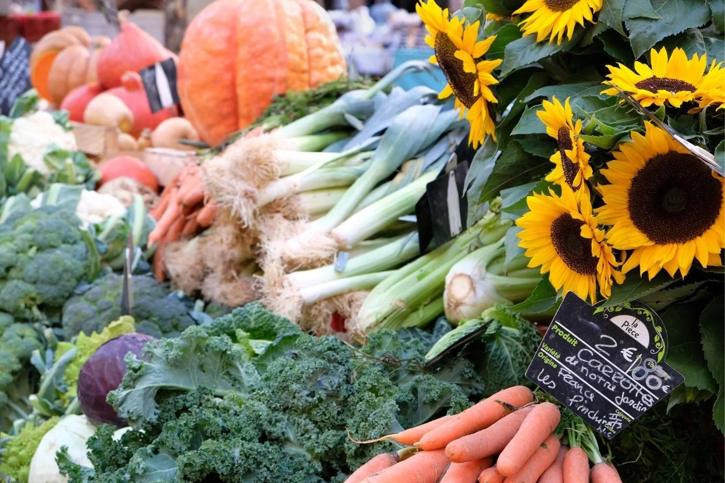 Fresh vegetables including carrots, leeks, broccoli, cauliflower, and cauliflower at a farmers market stand alongside bright yellow sunflowers.