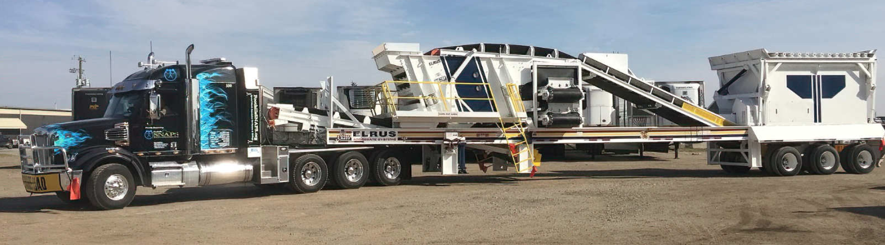 A large truck transporting heavy machinery, possibly a mobile manufacturing or processing unit, on a dirt surface under a cloudy sky.