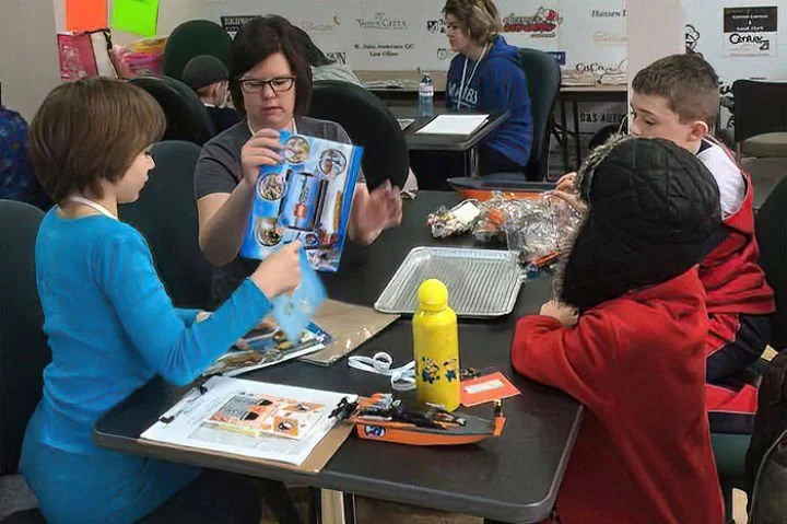 A woman and three children sit at a table with various craft supplies, including a yellow water bottle, plastic wrap, and snack items, in a busy indoor setting.