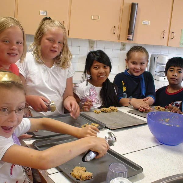 Five children in a kitchen preparing cookies, some are using scoops and others are arranging cookie dough on baking sheets, with a blue mixing bowl in front.