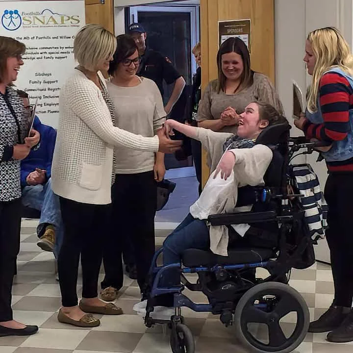 Group of women and a young woman in a wheelchair exchanging smiles and handshakes indoors.