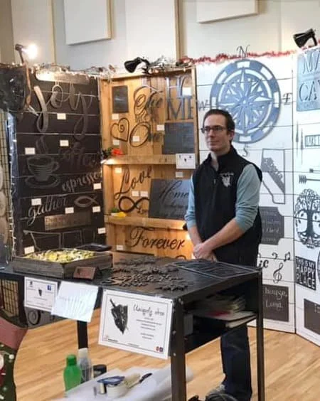 A man standing behind a black table at an indoor craft or home decor booth, with wall displays of wooden signs, black and white art, and decorative items, some with words like 'Love' and 'Forever.'