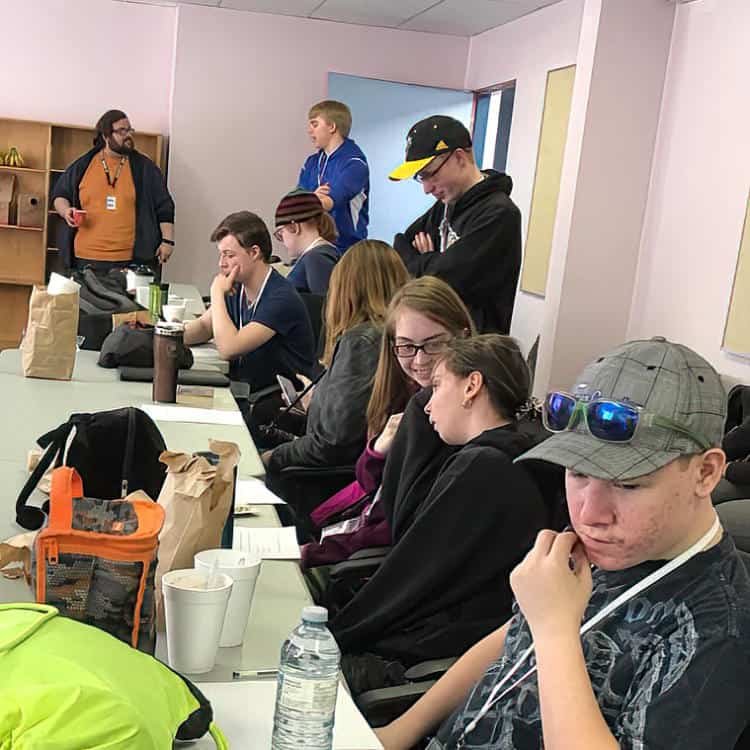 Group of teenagers sitting at a long conference table in a classroom or meeting room, some using smartphones, with bags, drinks, and notebooks on the table. Four people are standing in the background, one speaking and three listening.