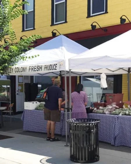 People at a farmers market booth selling fresh produce under a white canopy with a sign that reads 'Key Colony Fresh Produce' in front of a yellow building.