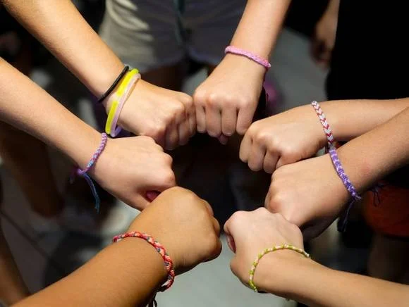 Six hands forming a circle, each wearing colorful braided bracelets.