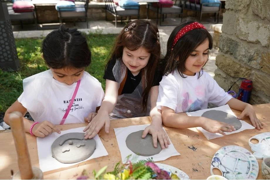Three girls working on clay molds at a table outdoors, with arts and crafts supplies nearby.
