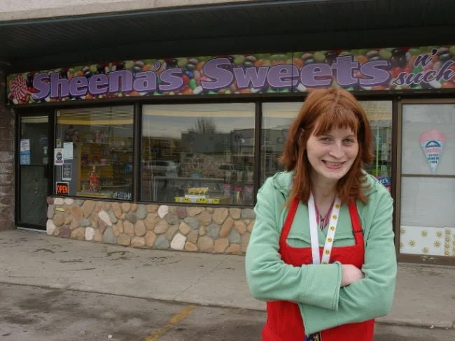 A young woman with red hair smiling outside a candy store named 'Sheena's Sweets.'