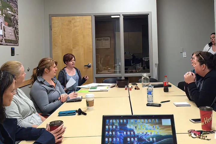 Group of people sitting around a conference table in a meeting room, with one woman standing and speaking.