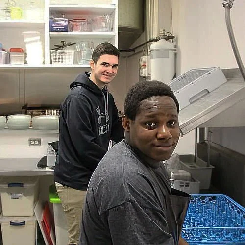 Two young men in a kitchen, one with dark skin and short curly hair, wearing a dark T-shirt, and the other with light skin and short dark hair, in a black hoodie, standing near a commercial kitchen sink and shelves with kitchen supplies.