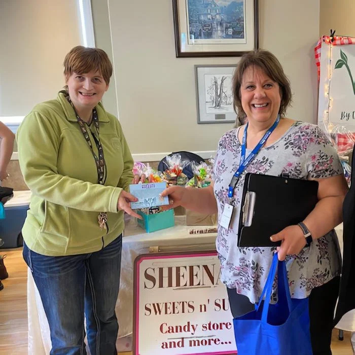 Two women smiling and exchanging a check inside a candy store. One woman wears a green jacket, and the other wears a floral top with a blue lanyard. There is a sign that says "SHEEN SWEETS n' S" and a table with candies and items in the background.