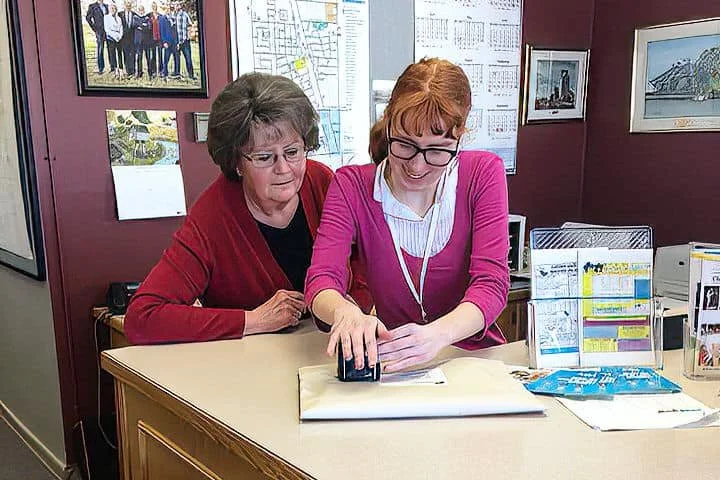 Two women working together at an office desk. The woman on the right, with red hair and glasses, is smiling and using a stamp, while the woman on the left, with short gray hair, watches closely. Behind them are framed artwork and documents on the wall.