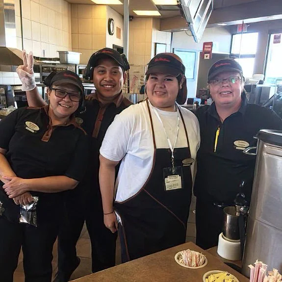 Four McDonald's employees posing inside a restaurant. They are smiling, with three women and one man, all wearing McDonald's uniforms. The woman in the center has a white shirt and black apron, while the others are in black shirts and hats.