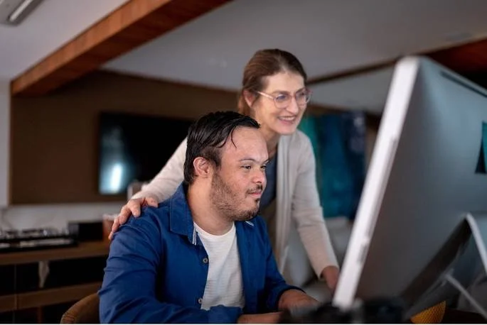 A man working on a computer with a woman standing behind him, smiling and looking at the screen in a modern office setting.