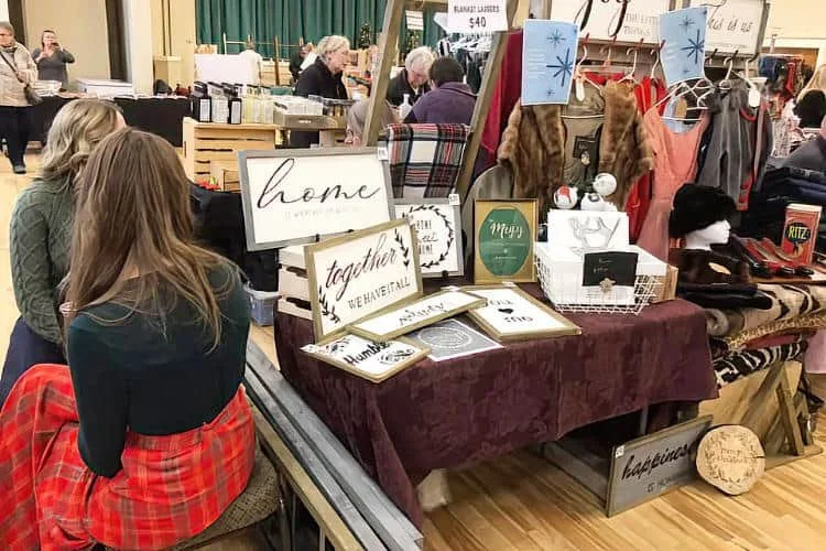 A table at a craft fair or market displaying home decor and gift items, including framed signs with words like "home" and "together," along with decorative accessories and clothing. People are shopping and conversing nearby.
