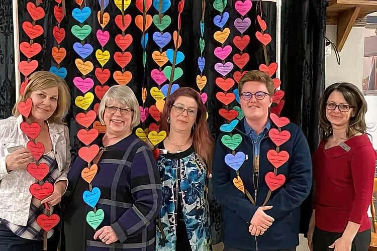 Five women standing in front of a colorful backdrop of hanging paper hearts in pink, red, orange, yellow, green, blue, and purple.