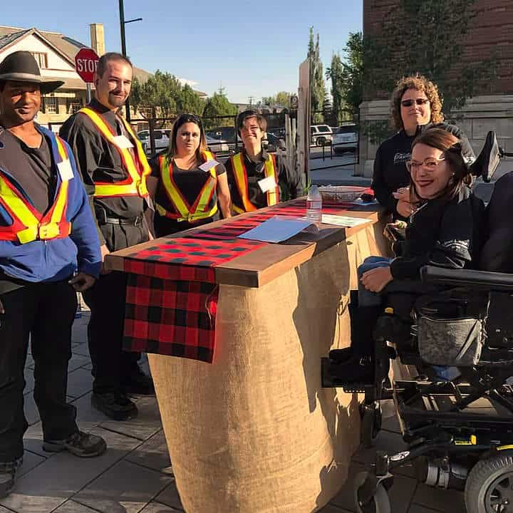 Group of six people, some wearing reflective safety vests, gathered around a table with a red and black checkered tablecloth outdoors in a city setting.
