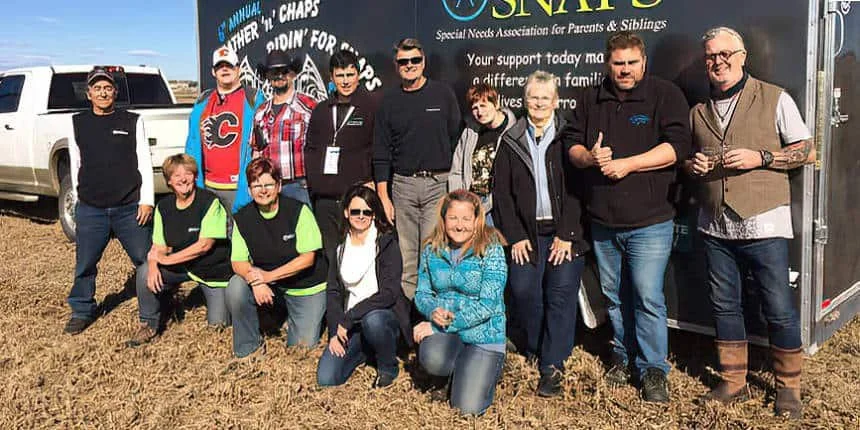 Group of people posing outdoors in front of a trailer with a sign for the Special Needs Association for Parents & Siblings, in a field on a sunny day.
