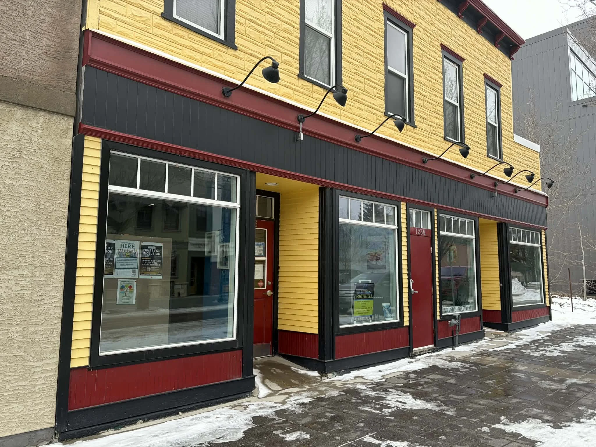 storefront with yellow and red exterior, large windows, three black outdoor sconce lights, and snow on sidewalk.