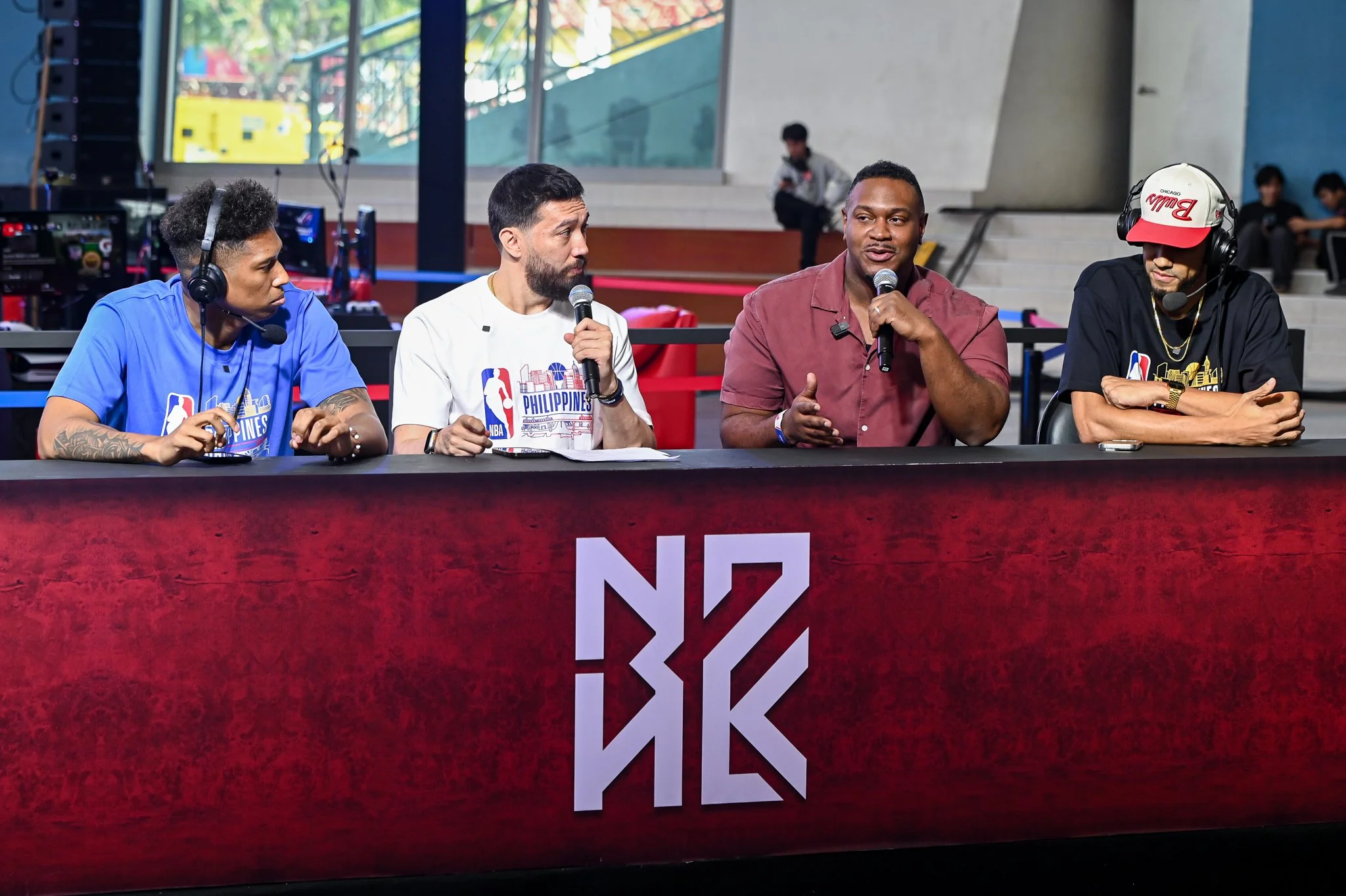 Four men sitting at a panel with microphones, wearing NBA and Philippines shirts, engaging in a discussion at an indoor venue with a red table and background spectators.