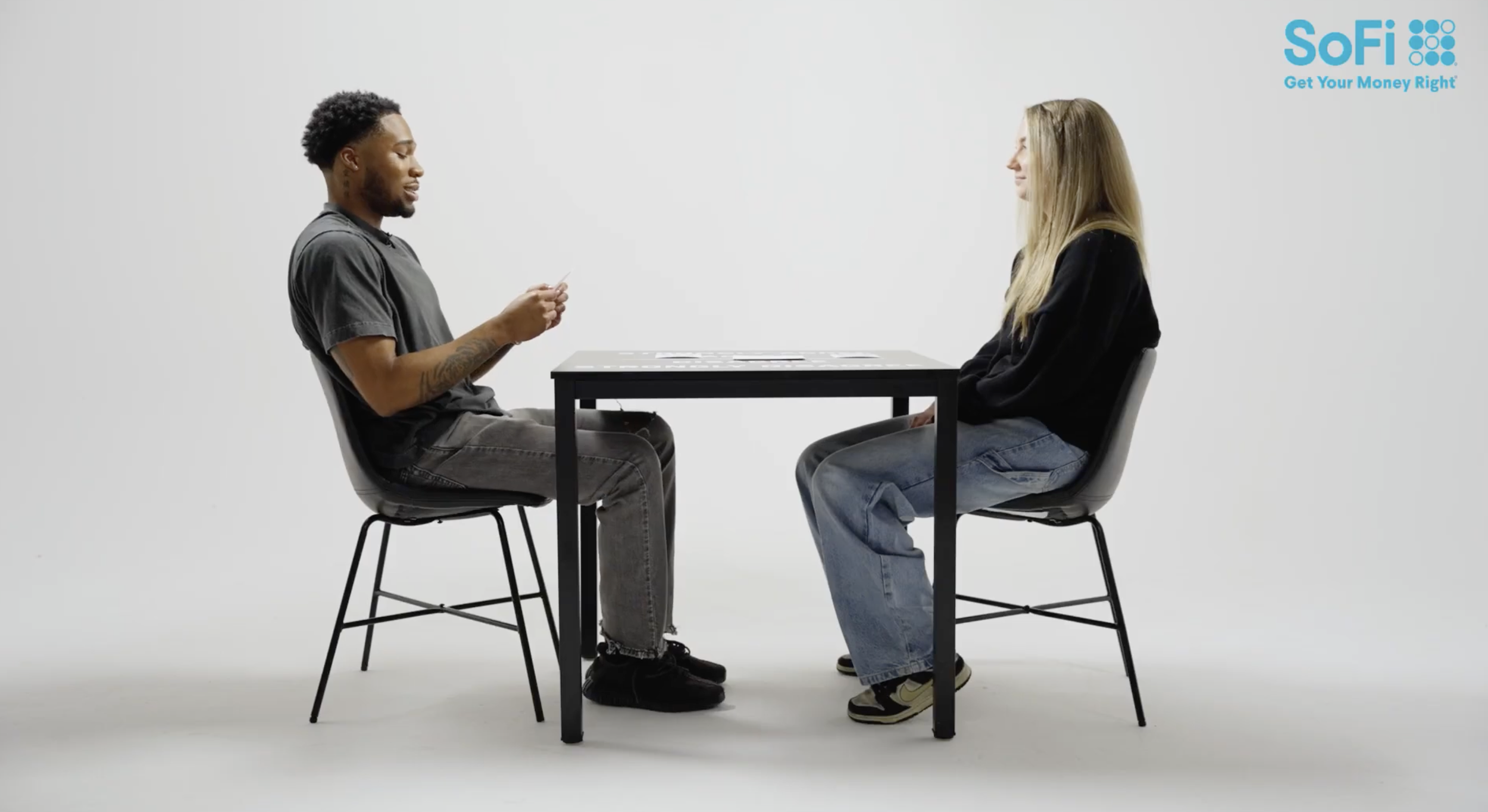 Two people sitting across a table from each other, the man on the left holding a card and the woman on the right sitting attentively. Both are seated on black chairs with a white background and a blue Sofi logo on the top right corner.
