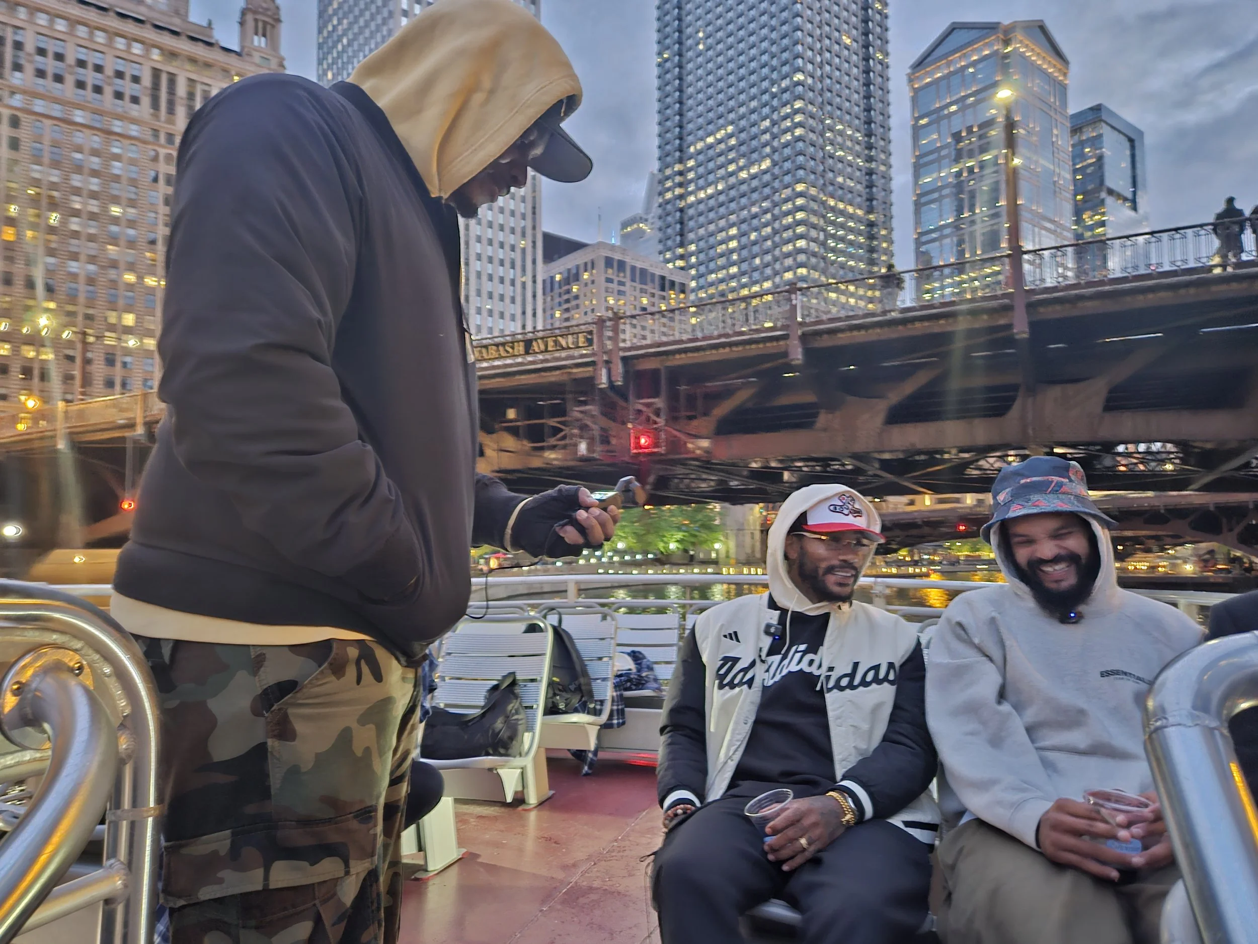 Three men hanging out outdoors in an urban area with tall buildings and a bridge in the background. Two men are sitting on a bench, smiling, while the third man is standing looking at his phone. All men are dressed casually, and it appears to be evening.