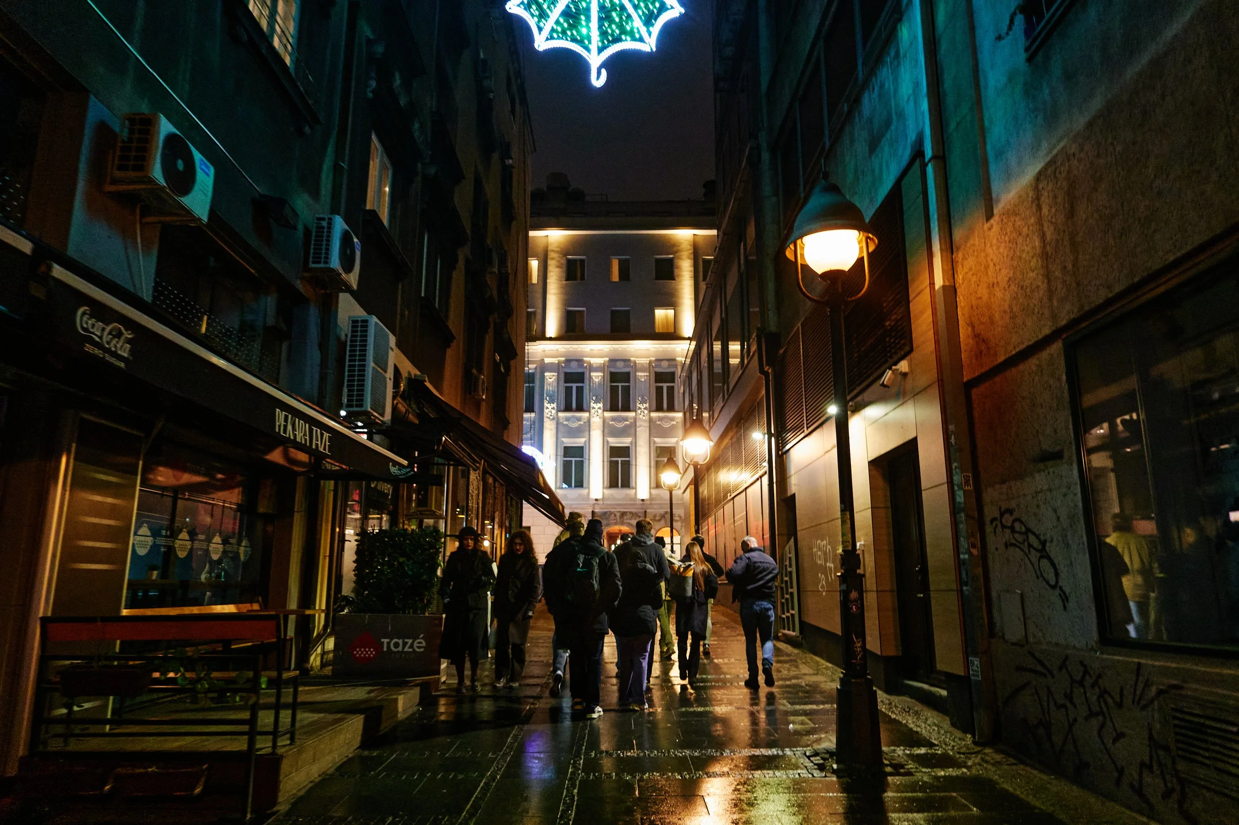 Night scene of a narrow street with pedestrians, illuminated by street lamps and decorative lights hanging above, with buildings on both sides.
