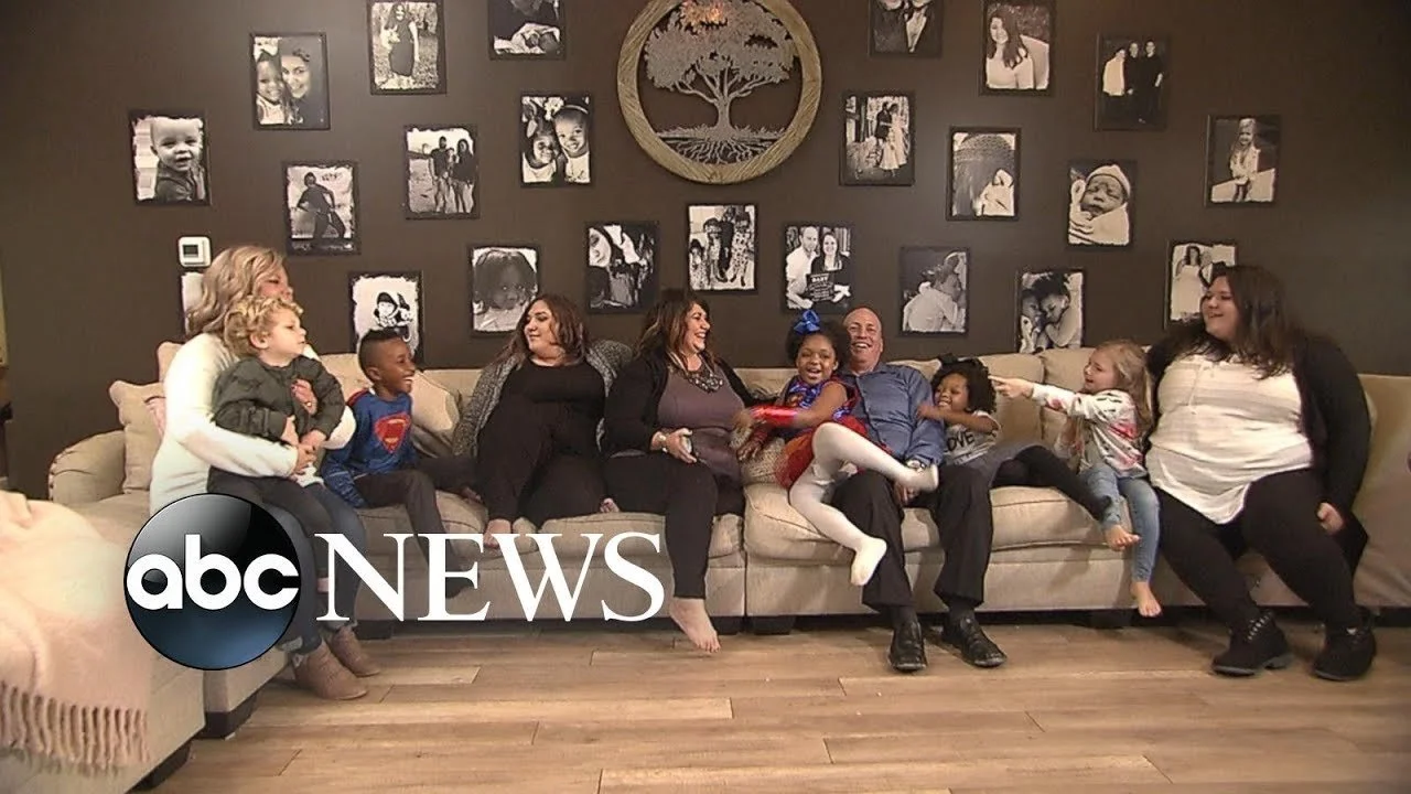 Family and friends sitting on a beige couch, smiling and playing together in a living room with a wall decorated with black and white photos and a wooden tree wall art.