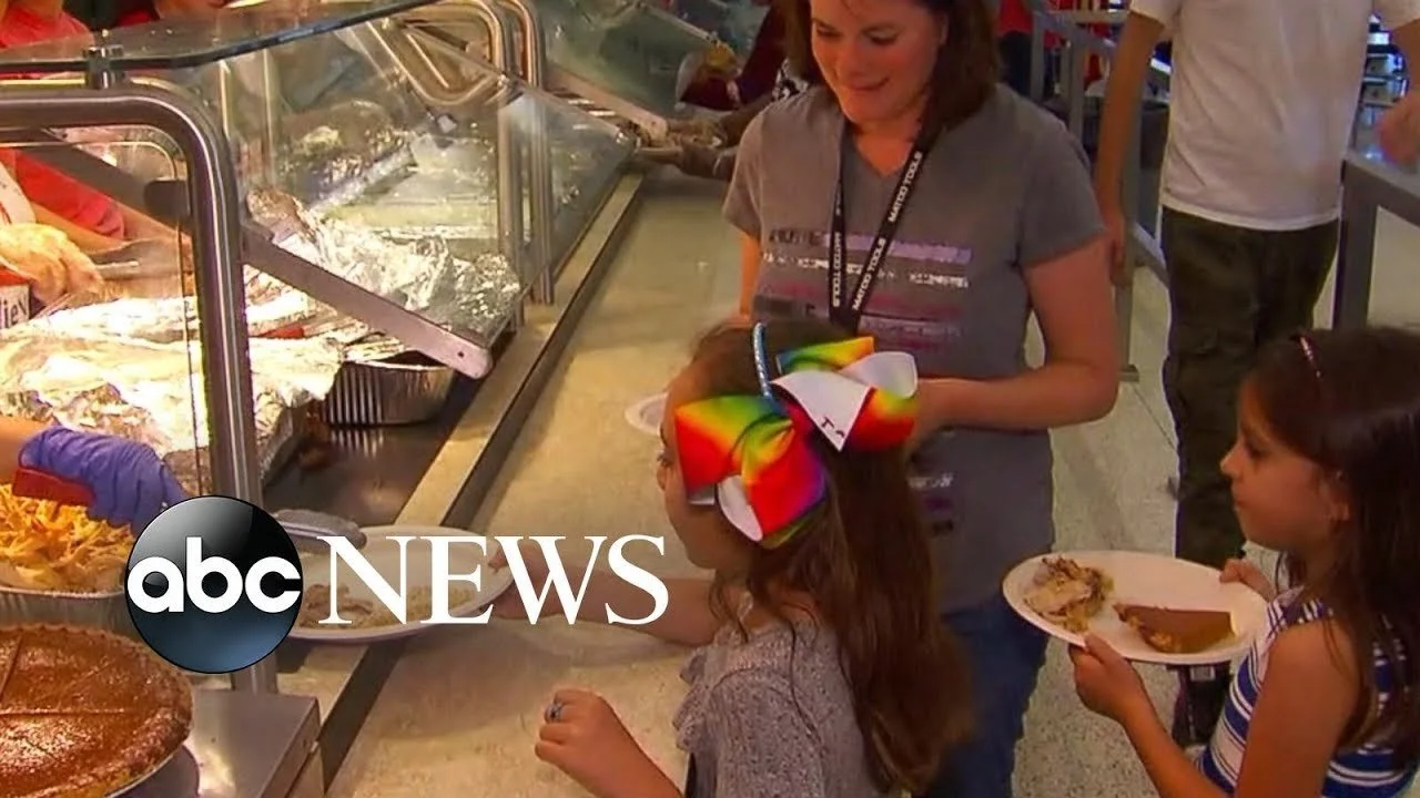 Two young girls and an adult woman at a buffet, selecting food from a glass display case. The girl on the right has long dark hair and a rainbow-colored bow, wearing a striped top. The woman is smiling and wearing a grey t-shirt with an ABC News logo
