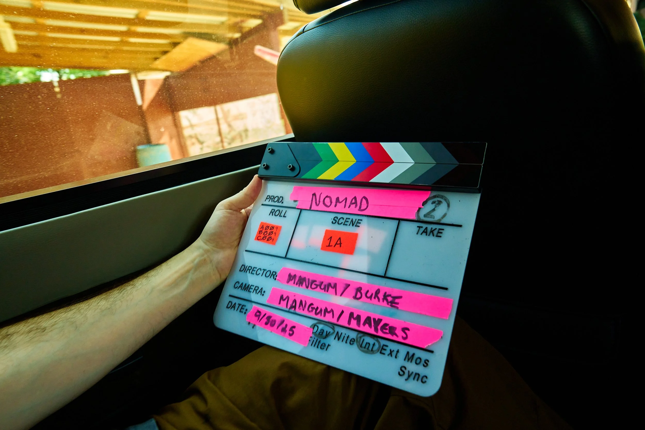 Clapperboard held by a person's hand inside a vehicle near a window, with greenery and wooden structures outside.