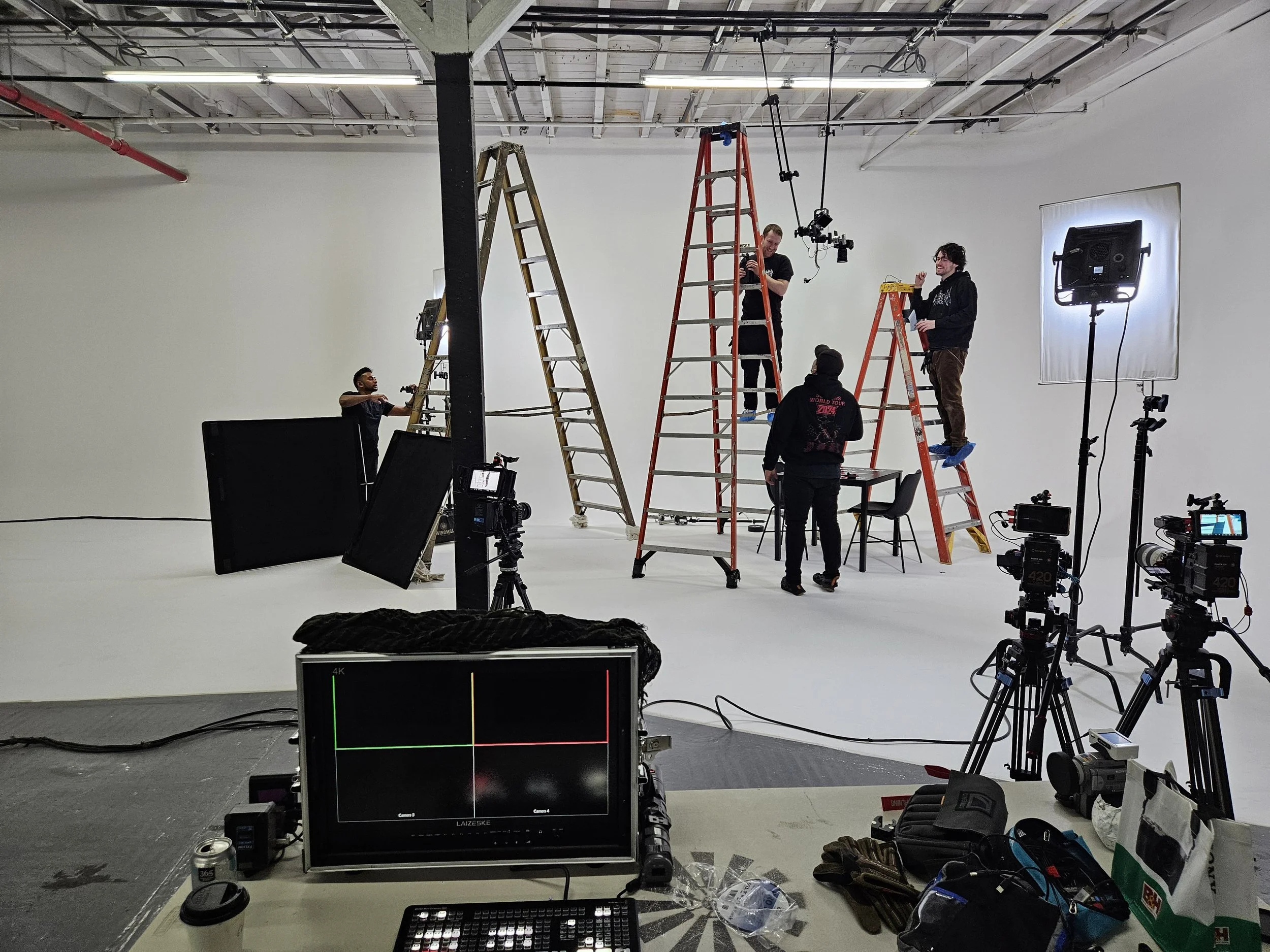 A film or photo shoot setup with crew members working on a white cyclorama. Several people are on ladders adjusting equipment, while others are standing around with cameras and lighting gear. Professional cameras, monitors, and lighting equipment are visible in the studio.